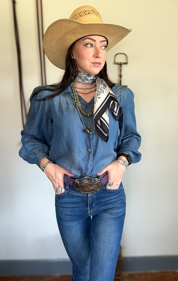 a girl wearing a blue button down shirt with turquoise jewelry on and a cowgirl hat. The outfit is western styled. 